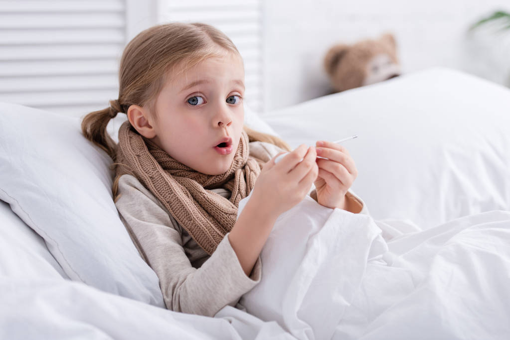 σοκαρισμένος άρρωστο παιδί με μαντήλι λαιμού ξαπλωμένη στο κρεβάτι και τον έλεγχο της θερμοκρασίας με θερμόμετρο στο σπίτι - Φωτογραφία, εικόνα