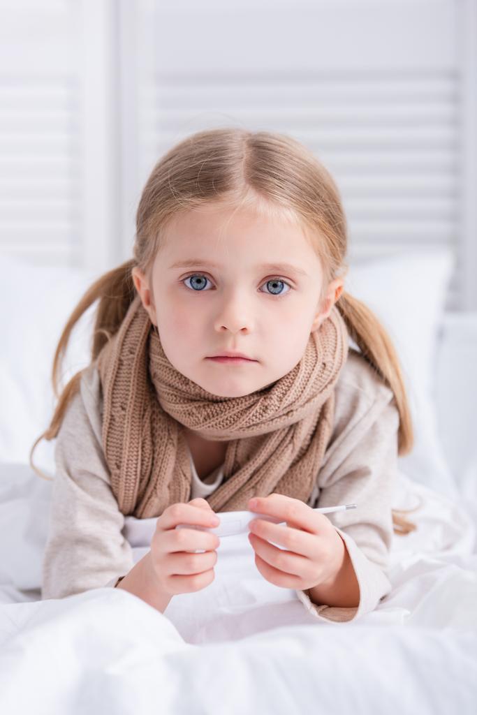 niño enfermo con bufanda sobre el cuello acostado en la cama, sosteniendo el termómetro y mirando a la cámara en casa
 - Foto, Imagen