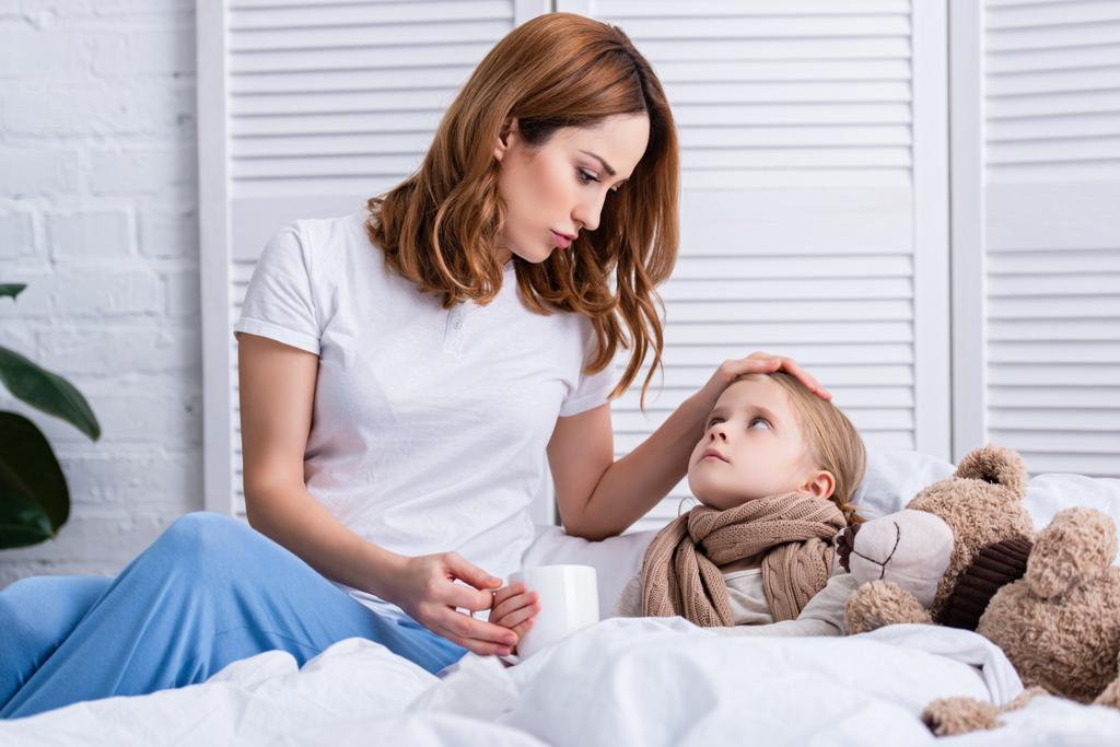 mother taking care of sick daughter in bedroom and giving her cup of tea, looking at each other - Photo, Image
