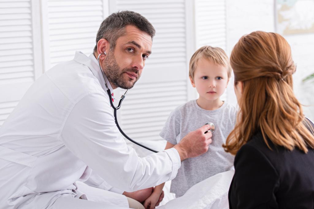 pediatrist in white coat examining sick boy with stethoscope and looking at camera at home - Photo, Image