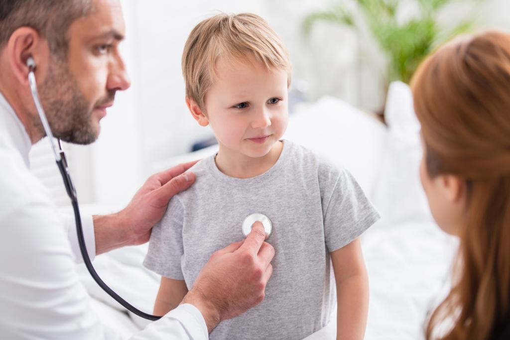 selective focus of pediatrist in white coat examining sick boy with stethoscope - Photo, Image