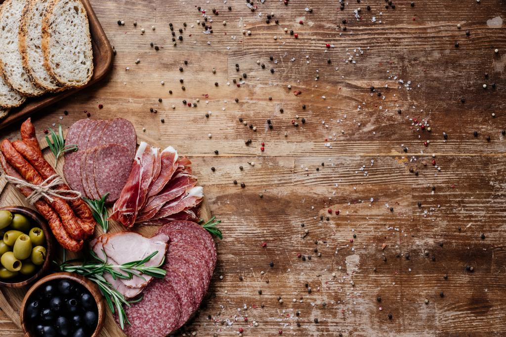 bovenaanzicht van snijplanken met heerlijke salami, gerookte worst, olijven en brood op houten tafel met verspreide peperkorrels - Foto, afbeelding
