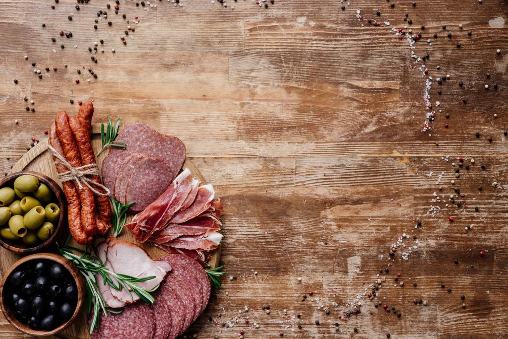 Rundschneidebrett mit schmackhaften Salami, Schinken und Oliven in Schalen auf Holztisch mit verstreuten Gewürzen - Foto, Bild