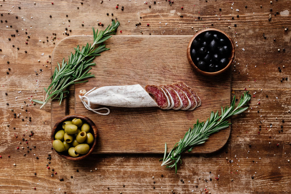 vue du dessus de la planche à découper avec romarin, olives noires et vertes dans deux bols et délicieux salami tranché sur une table en bois avec des épices éparses
 - Photo, image