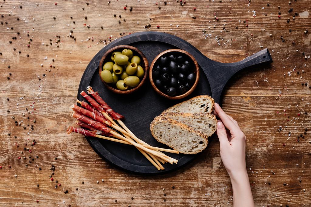 cropped view of  woman putting bread on round cutting board with olives, breadsticks and prosciutto on wooden table - Photo, Image