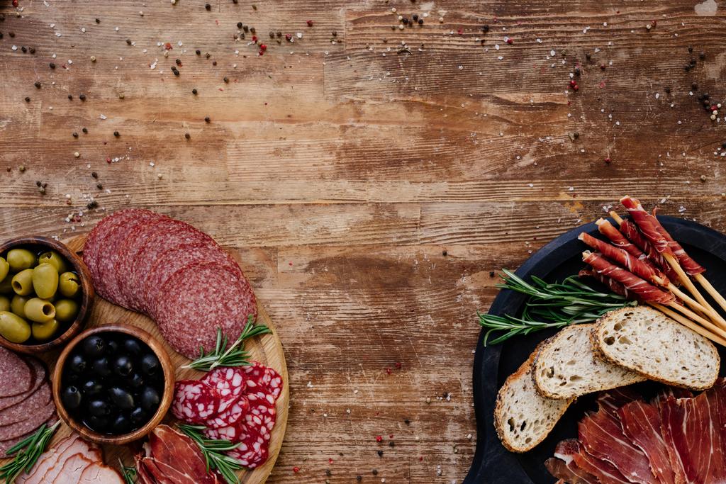 top view of cutting boards with olives, breadsticks, prosciutto, salami, bread and herbs on wooden table - Photo, Image