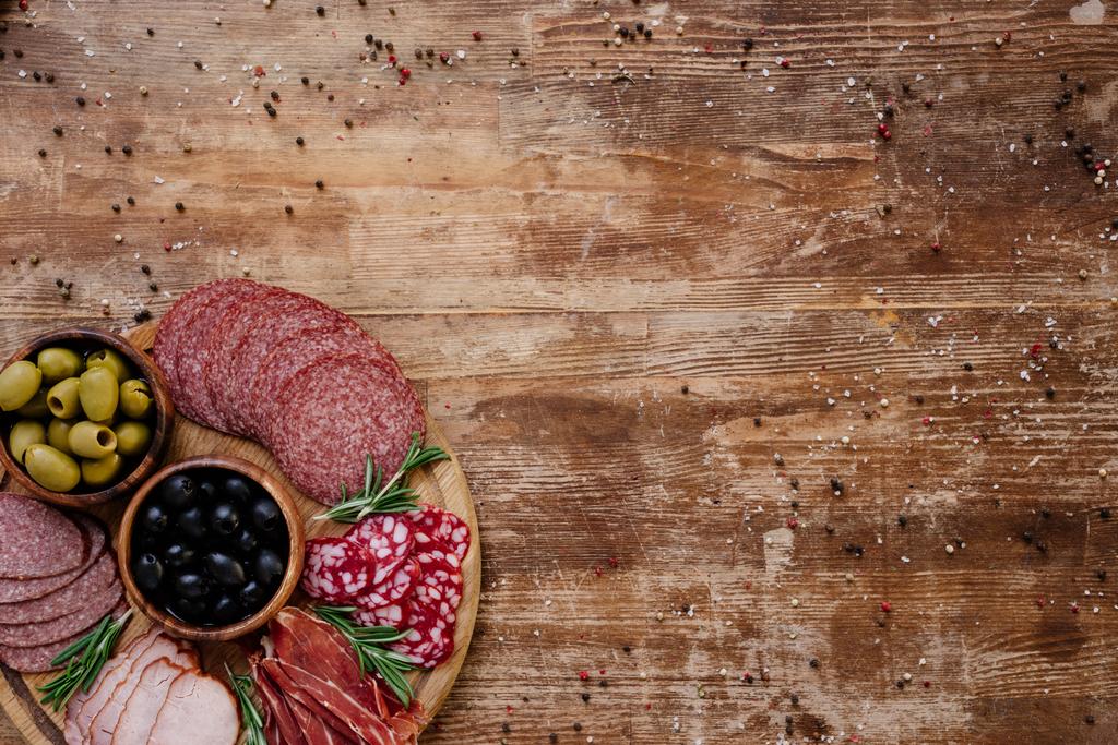 Draufsicht auf Schneidebrett mit Oliven, Stockbrot, köstlichem Prosciutto, Salami und Kräutern auf Holztisch mit verstreuten Gewürzen - Foto, Bild