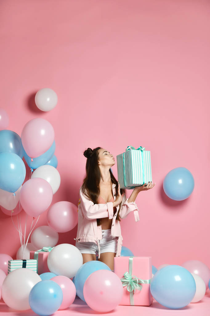 Portrait of young woman in birthday party holding gift box with gift present on pastel pink background with colorful air baloons.  - Photo, Image
