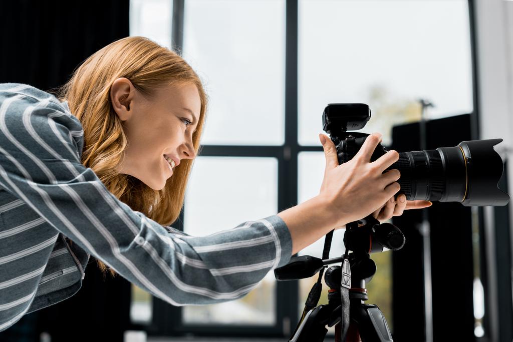 Zijaanzicht van lachende jonge vrouwelijke fotograaf werkt met professionele fotocamera in studio    - Foto, afbeelding