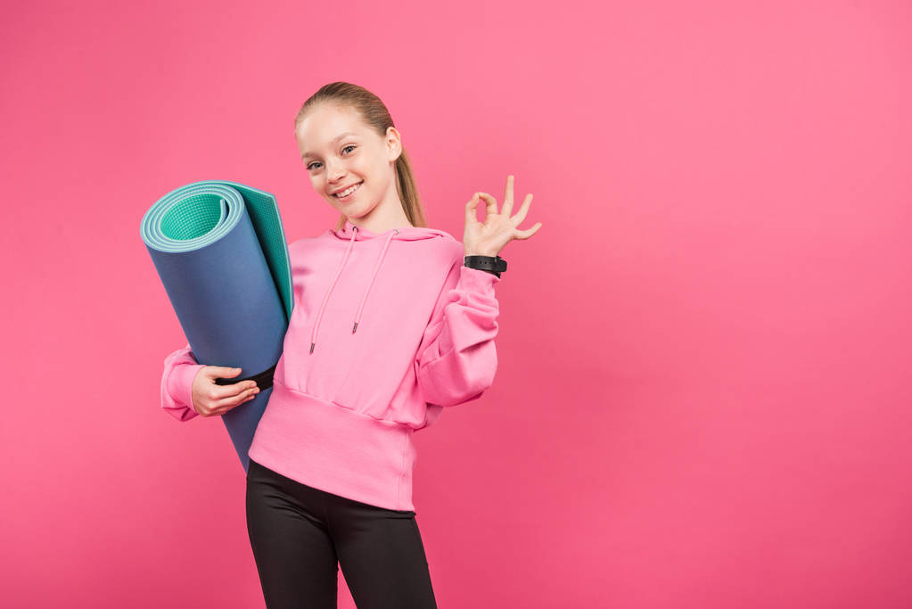 счастливый ребенок, держащий фитнес-коврик и показывающий знак ОК, изолированный на розовом
 - Фото, изображение