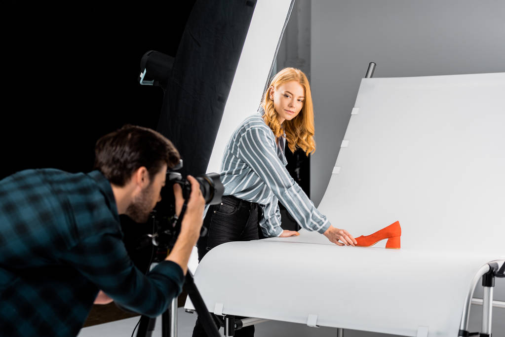 αρσενικό φωτογράφος εργάζεται με φωτογραφική μηχανή, ενώ συνάδελφος τακτοποίηση παπουτσιών στο studio  - Φωτογραφία, εικόνα