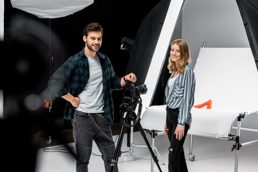jóvenes fotógrafos felices de pie cerca de equipo fotográfico profesional y sonriendo a la cámara en el estudio
  - Foto, Imagen