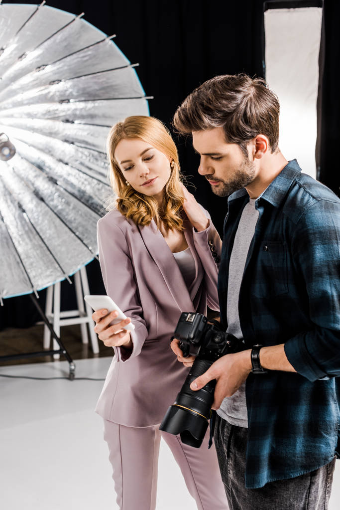 φωτογράφος με κάμερα και όμορφο γυναικείο μοντέλο χρησιμοποιώντας smartphone σε φωτογραφία στούντιο  - Φωτογραφία, εικόνα