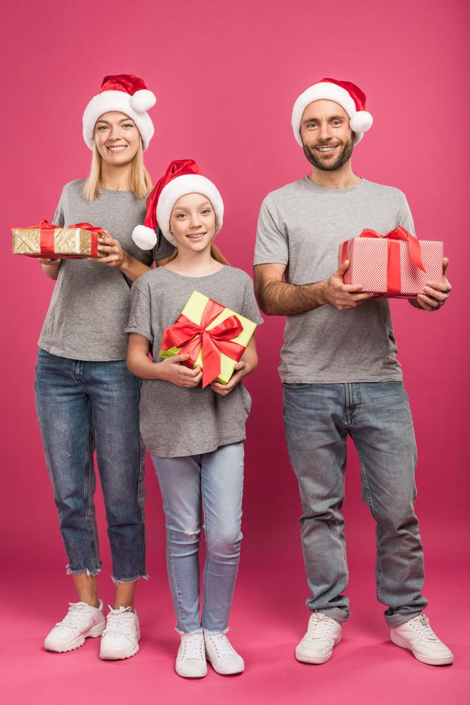 щаслива сім'я в капелюхах Санти, що тримає різдвяні подарункові коробки на рожевому
 - Фото, зображення