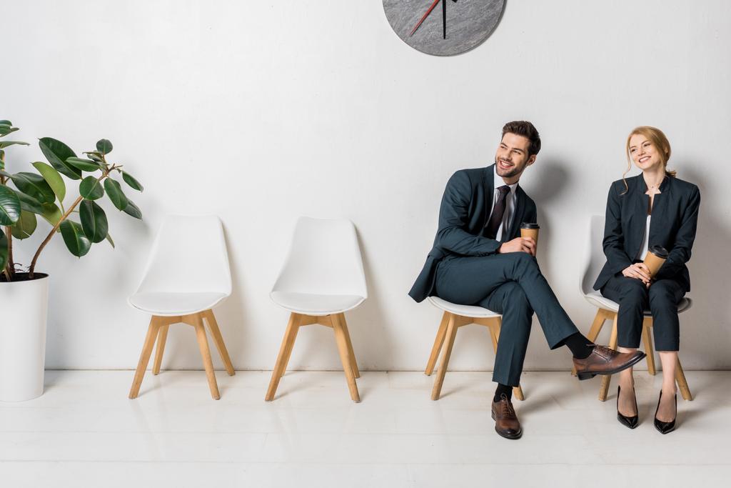 χαμογελώντας νέοι επιχειρηματίες κρατώντας χαρτί Κύπελλα και αναζητούν μακριά ενώ περιμένουν στις καρέκλες στη γραμμή - Φωτογραφία, εικόνα