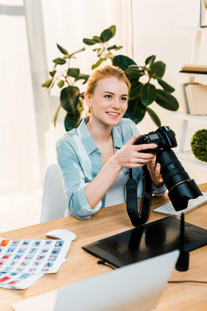 όμορφη χαμογελαστός νεαρός φωτογράφος κρατώντας κάμερα ενώ εργαζόταν στο γραφείο  - Φωτογραφία, εικόνα