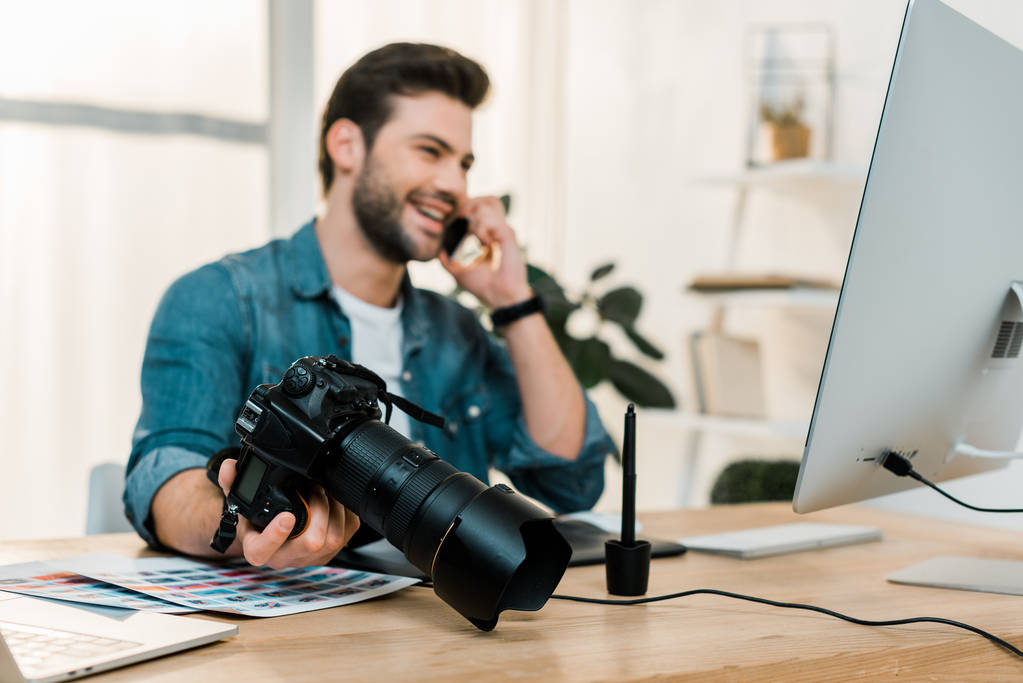 jovem fotógrafo feliz segurando câmera e falando por smartphone na mesa de trabalho
 - Foto, Imagem