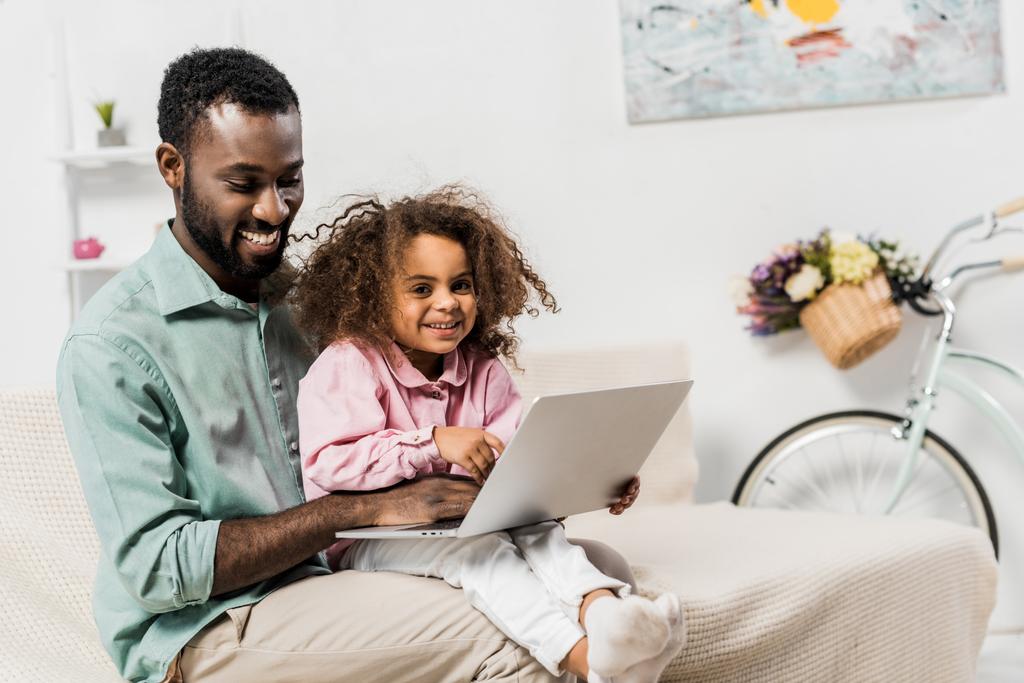 padre afroamericano y adorable niño sentado en el sofá y el uso de ordenador portátil
 - Foto, Imagen