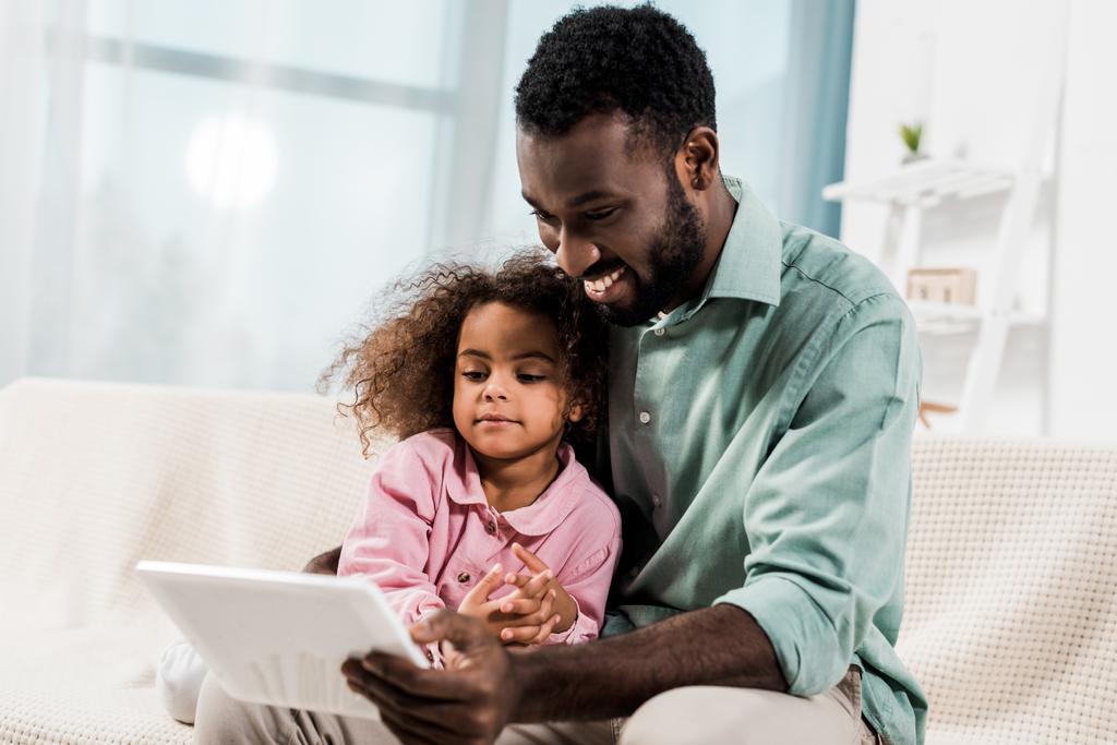 sonriente afroamericano padre y niño usando tableta digital mientras está sentado en el sofá
 - Foto, imagen