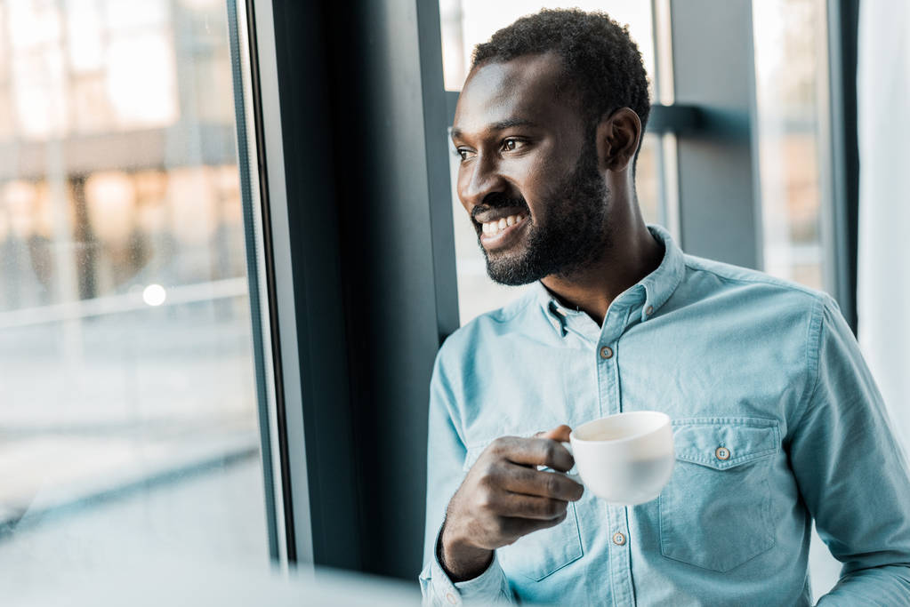 窓の外を見ながら一杯のコーヒーを保持しているハンサムなアフリカ系アメリカ人 - 写真・画像