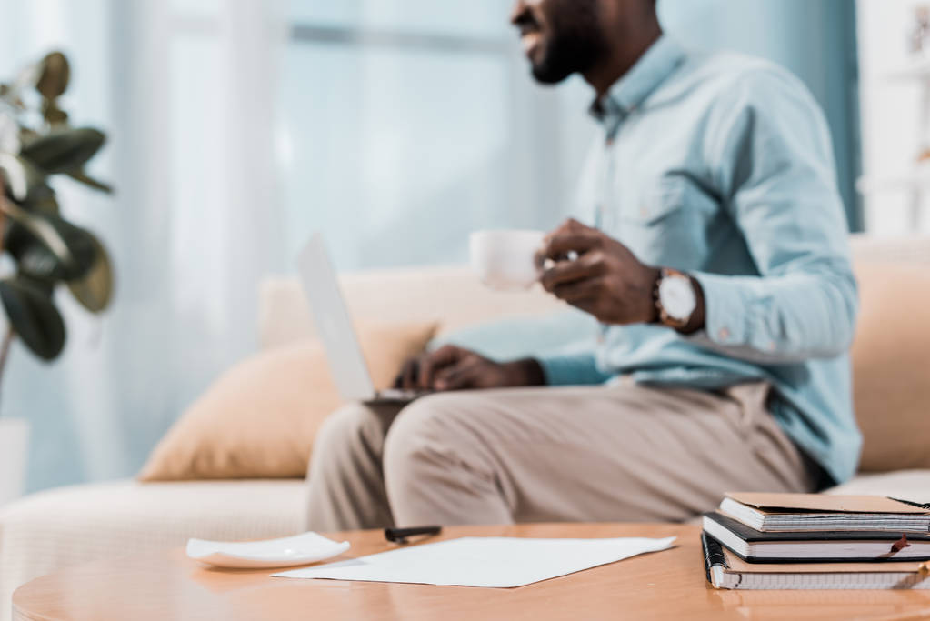 vista cortada de Africano americano freelancer sentado no sofá com laptop e beber café
 - Foto, Imagem
