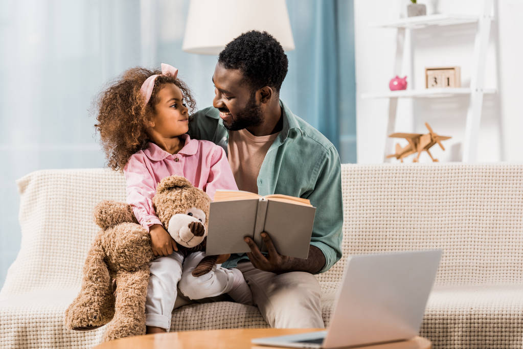 papá afroamericano leyendo con su hija y mirándose en la sala de estar
 - Foto, Imagen