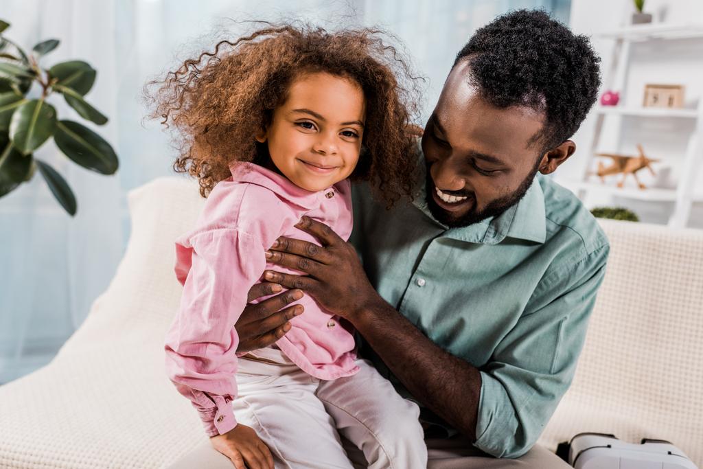 Afrikanisch-amerikanischer Vater hält lächelnde Tochter auf Knien im Wohnzimmer - Foto, Bild