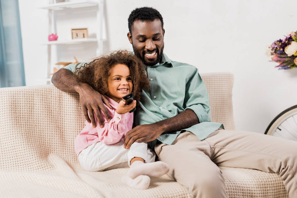 Αφρικανική αμερικανική μπαμπάς αγκαλιάζει κόρη ενώ παρακολουθείτε τηλεόραση στο σαλόνι - Φωτογραφία, εικόνα