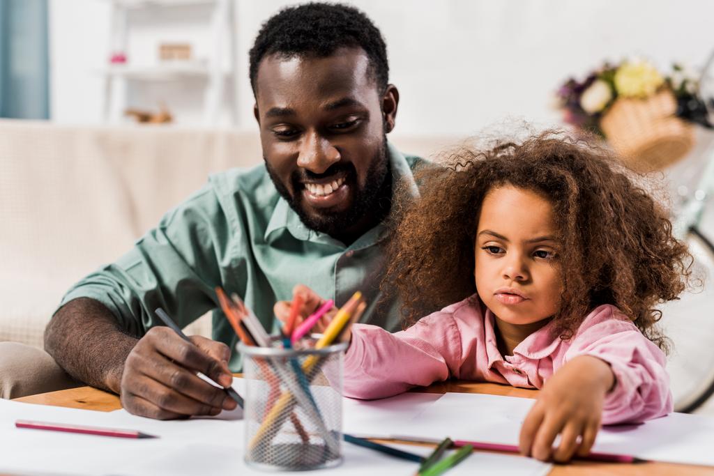 крупним планом вид на афроамериканського батька, який допомагає доньці з малюнком у вітальні
 - Фото, зображення
