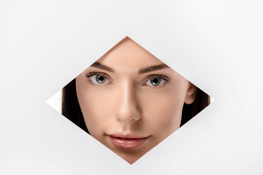 beautiful young woman looking at camera through rhombus shaped hole on white - Photo, Image