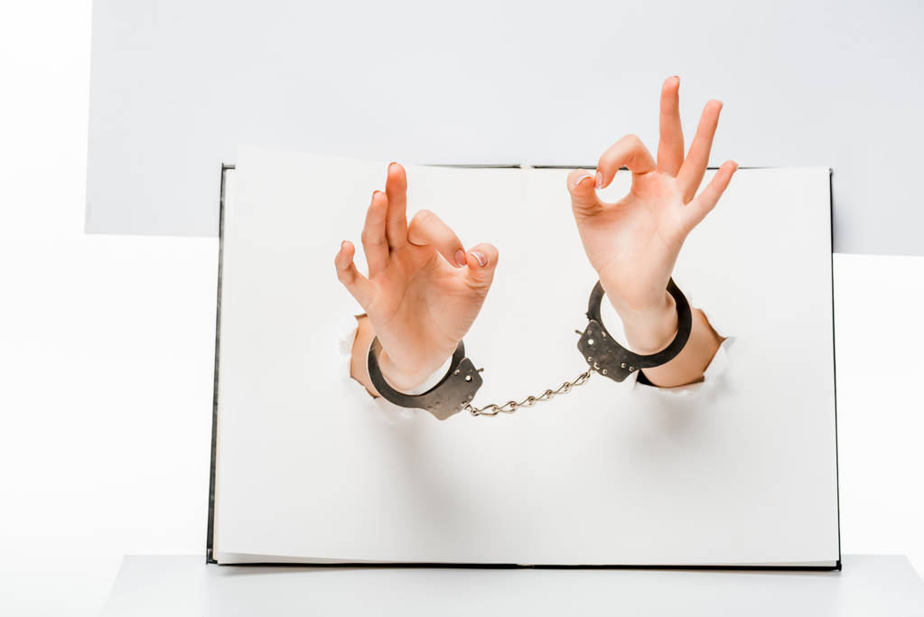 partial view of female hands in handcuffs showing ok sign through holes on white  - Photo, Image