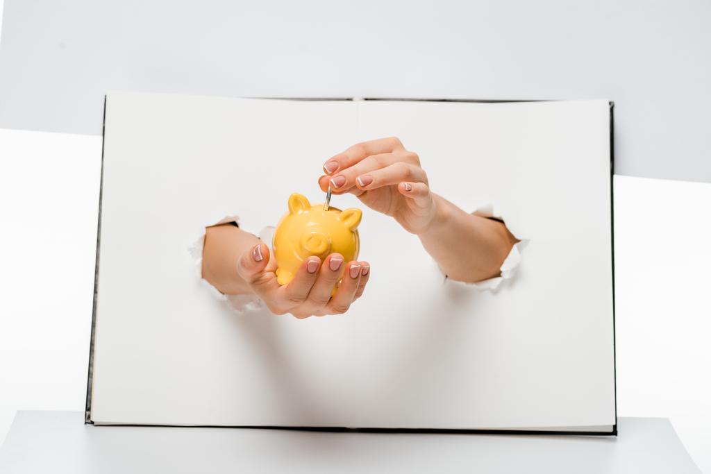 abgeschnittene Aufnahme einer Frau, die Münze durch Löcher auf Weiß in ein gelbes Sparschwein steckt  - Foto, Bild