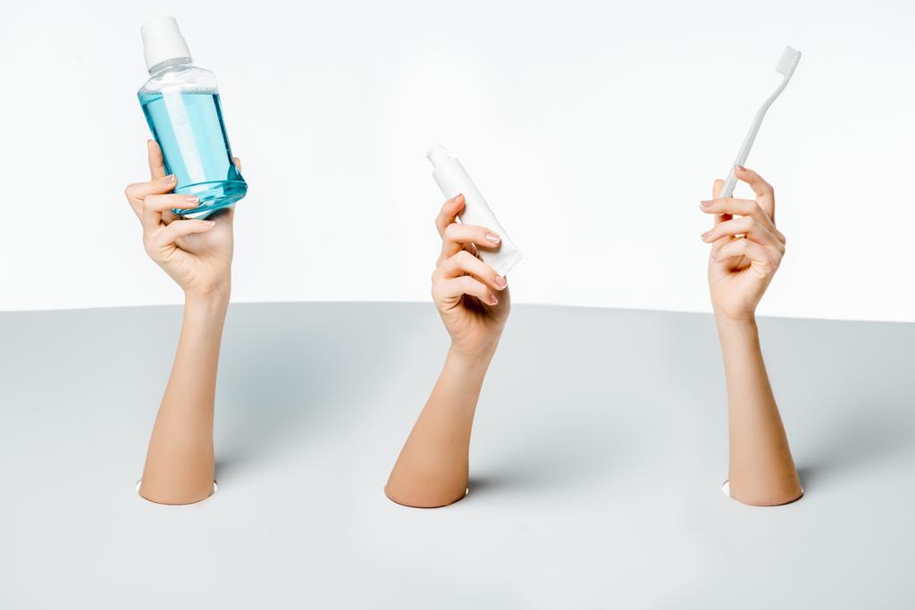 περικοπεί shot των γυναικών που κατέχουν οδοντόβουρτσα, οδοντόπαστα και στοματικό διάλυμα μέσα από τρύπες σε λευκό  - Φωτογραφία, εικόνα