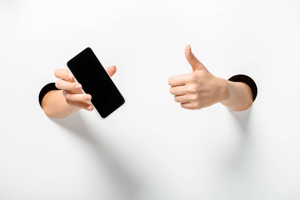 обрізане зображення жінки, що тримає смартфон з порожнім екраном і показує великий палець вгору через отвори на білому
 - Фото, зображення