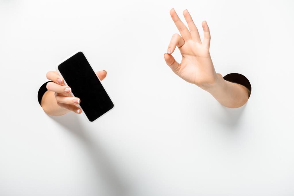przycięte zdjęcie kobiety, trzymając smartfon z pusty ekran i pokazuje gest porządku przez otwory na białym - Zdjęcie, obraz