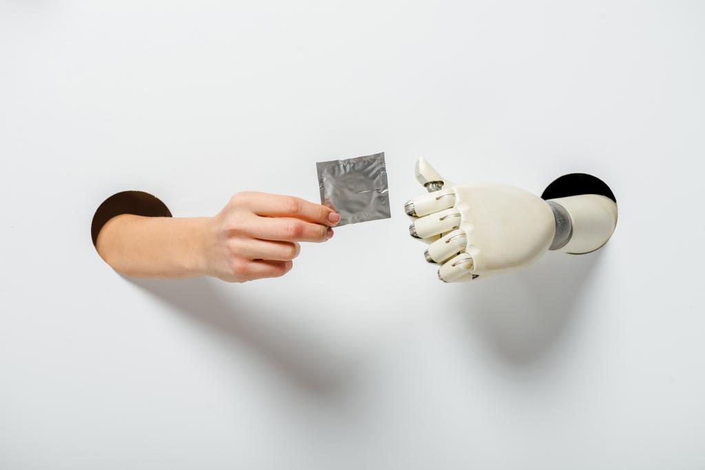 περικομμένη εικόνα μιας γυναίκας κι ενός ρομπότ κρατώντας προφυλακτικό μέσα από τρύπες σε λευκό - Φωτογραφία, εικόνα
