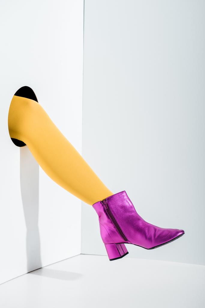 oříznutý obraz ženy ukazující nohy v zářivě žluté punčocháče a ultra fialové boty v díře na bílém - Fotografie, Obrázek