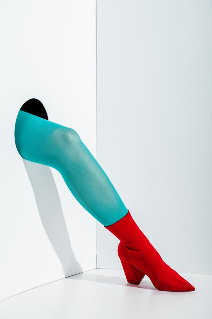 oříznutý obraz dívka ukazuje nohy v stylové tyrkysové punčocháče a červené boty v díře na bílém - Fotografie, Obrázek