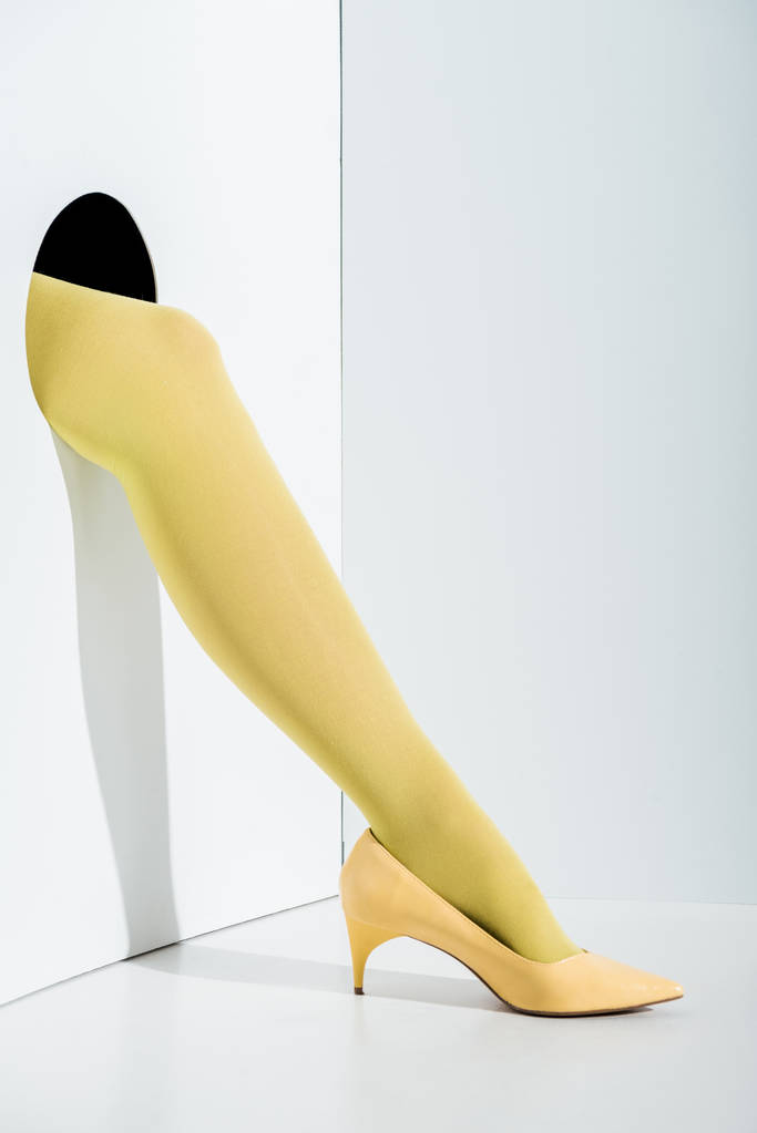 περικομμένη εικόνα κορίτσι δείχνει το πόδι σε κίτρινο καλσόν και ψηλοτάκουνα σε τρύπα στο λευκό - Φωτογραφία, εικόνα