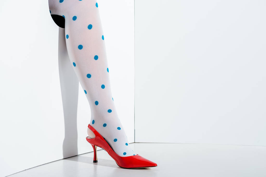 Ausschnittbild eines Mädchens, das Bein in weißen Strumpfhosen mit blauen Punkten und rotem High Heel im Loch auf weißem Grund zeigt - Foto, Bild