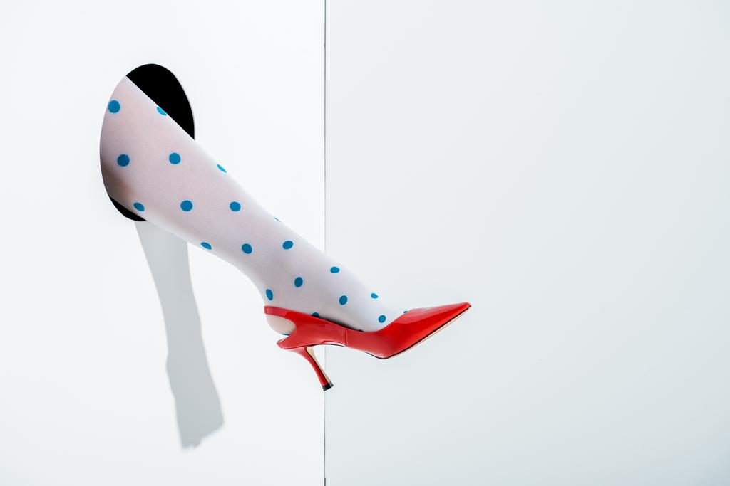 oříznutý obraz ženy ukazující nohy v bílých punčochách s modrými tečkami a červená vysoký podpatek v díře na bílém - Fotografie, Obrázek