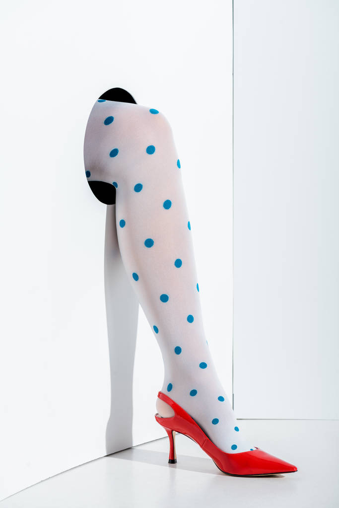 Ausschnittbild eines Mädchens, das Bein in stilvollen weißen Strumpfhosen mit blauen Punkten und rotem High Heel im Loch auf weißem Grund zeigt - Foto, Bild