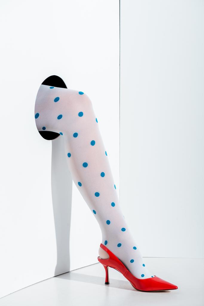 oříznutý obraz dívka ukazuje nohy v módní bílé punčocháče s modrými tečkami a červená vysoký podpatek v díře na bílém - Fotografie, Obrázek