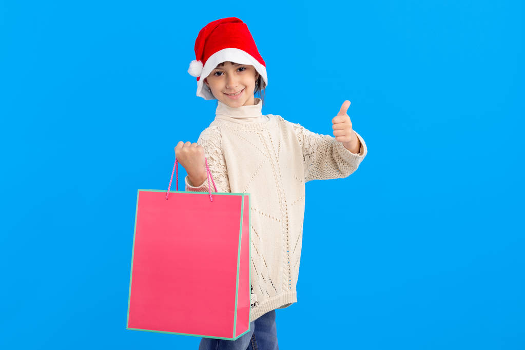 ¡Como! Hermosa chica en una chaqueta de invierno y sombrero de Santa con una bolsa de regalo rosa sobre un fondo azul. Pulgares hacia arriba - buenas compras para Navidad y Año Nuevo
. - Foto, Imagen