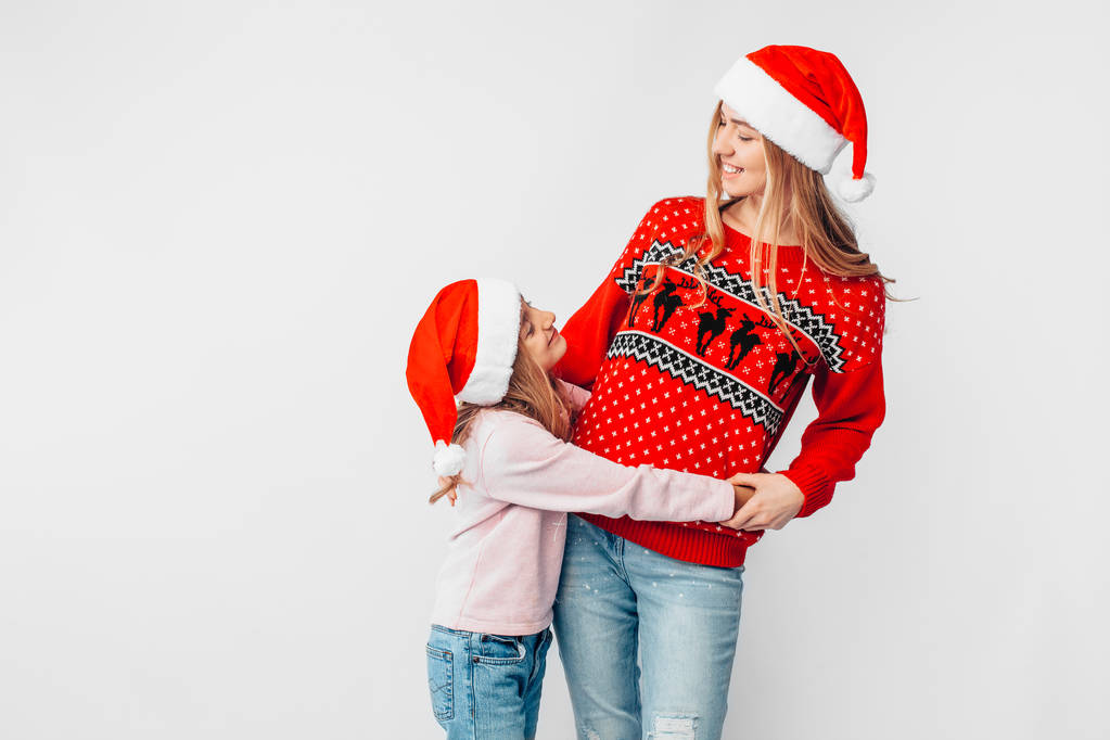 Mutter und Tochter tragen Weihnachtsmannhüte, und in Weihnachtspullis umarmen sie sich. auf weißem Hintergrund. das Konzept einer starken Familie. - Foto, Bild