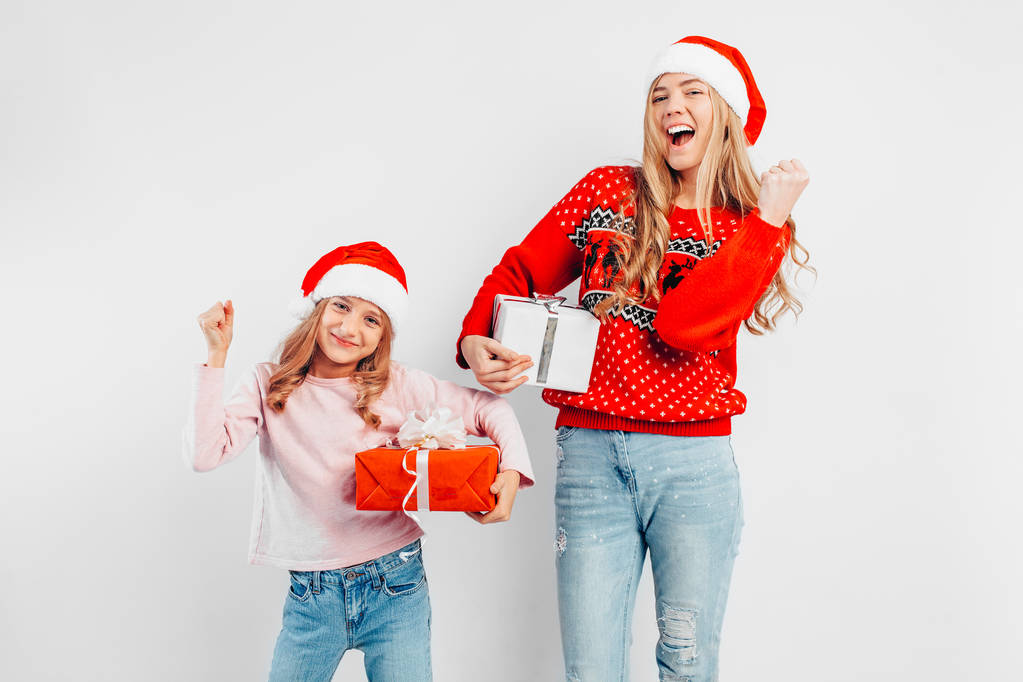 Schockierte Mutter und Tochter feiern das neue Jahr in Weihnachtsmützen und Pullovern mit Geschenken in der Hand auf weißem Hintergrund. - Foto, Bild