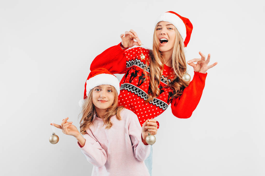 Мама и дочь носят шляпы Санта-Клауса, в новогодних свитерах, празднуют Новый год, играя с рождественскими шарами. На белом фоне
. - Фото, изображение