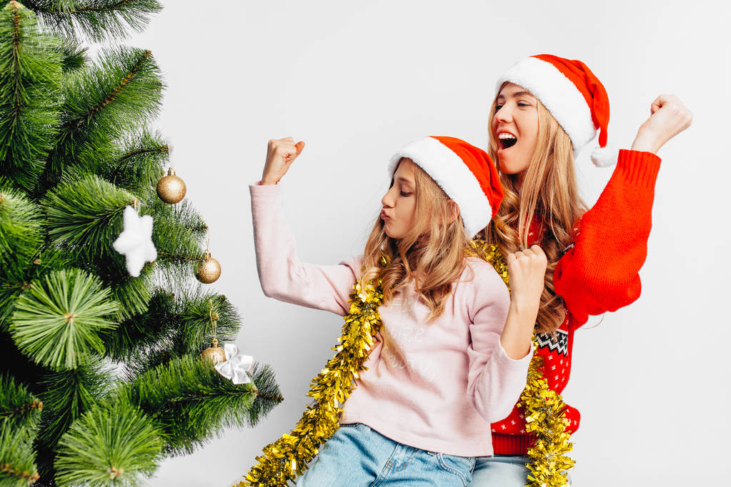 Felice mamma e figlia, in cappelli di Babbo Natale, seduti vicino all'albero di Natale, gioire, mostrando il gesto della vittoria, su sfondo bianco
. - Foto, immagini
