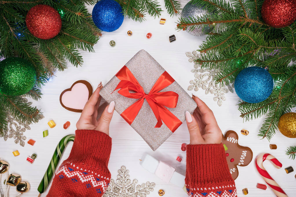 Kéz meleg pulóver gazdaság ajándék ezüst papírt a piros orr. Fából készült fehér asztal, karácsonyi dekoráció - fenyő ágak karácsonyi bálok, mézeskalács, nyalókák. - Fotó, kép