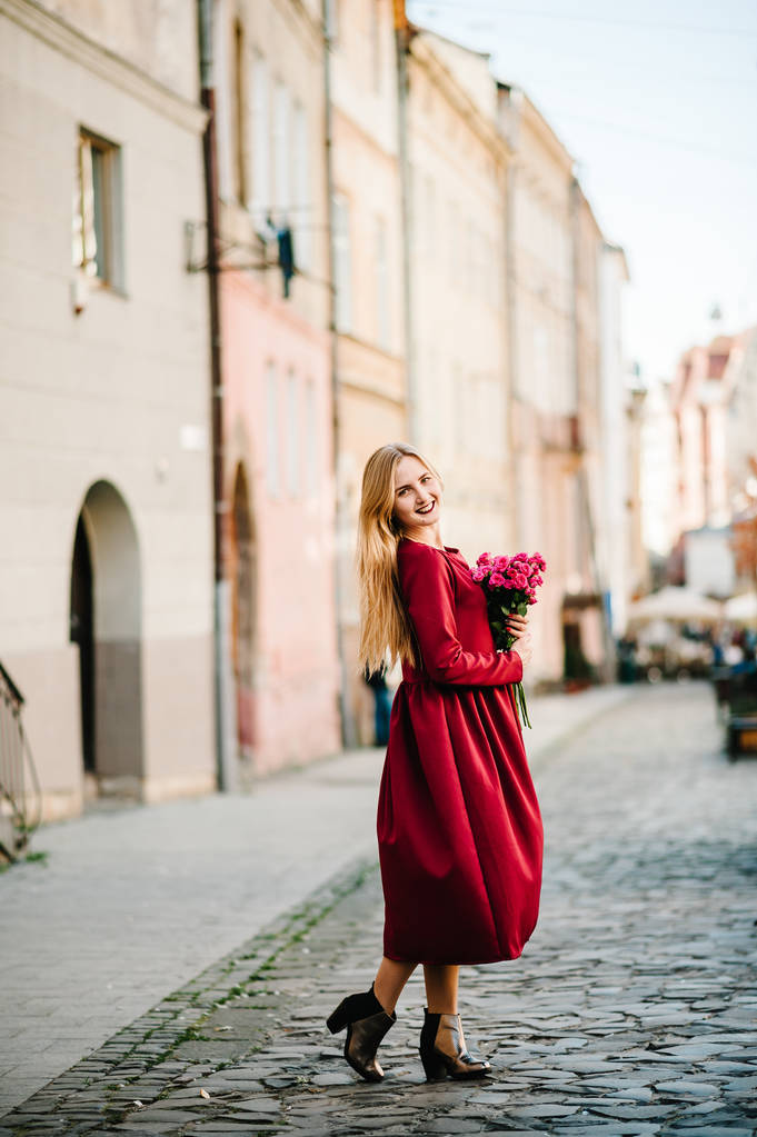 Porträt einer jungen, stilvollen Frau in rotem Kleid, die mit einem Rosenstrauß in der Altstadt posiert - Foto, Bild